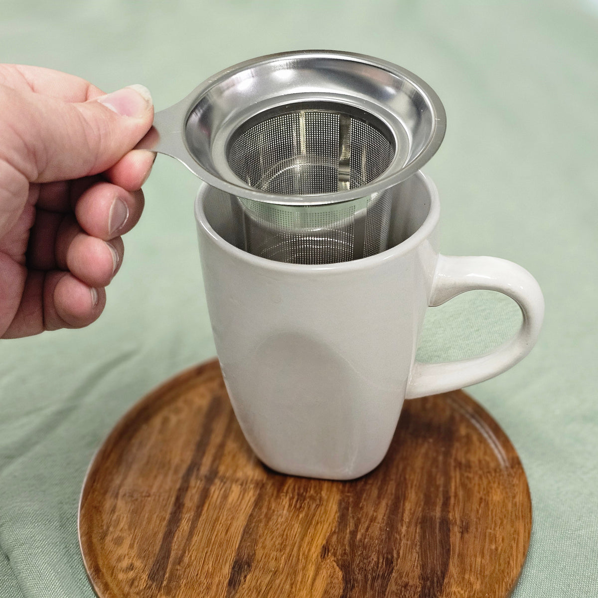 A hand holds a stainless steel tea infuser over a white ceramic mug, which sits on a round wooden coaster over a light green fabric surface.