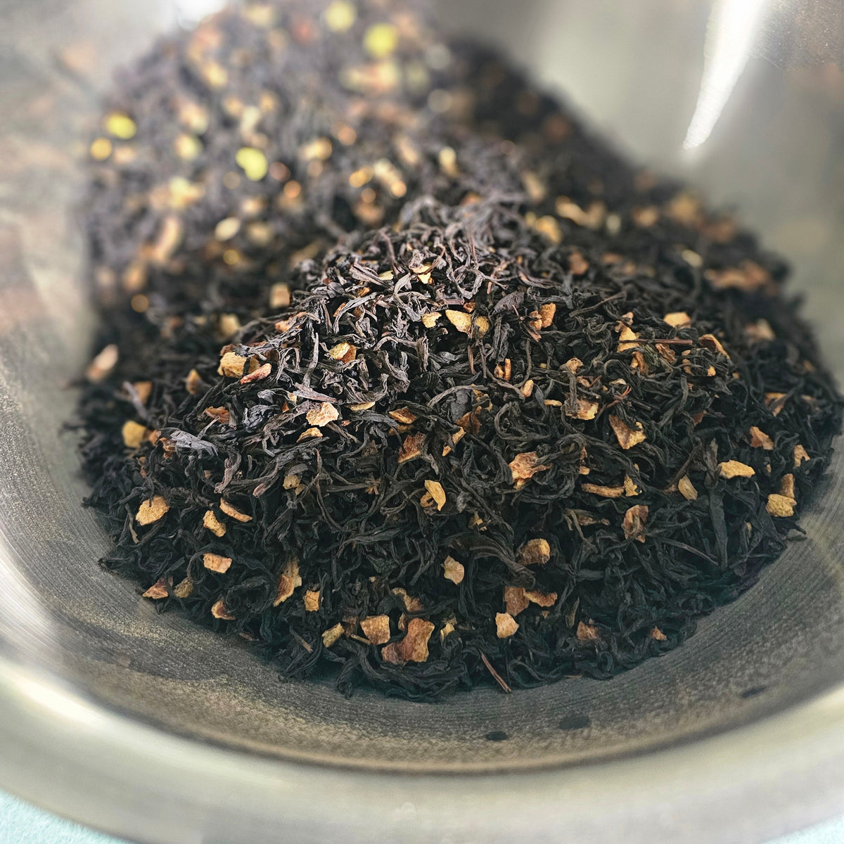 A close-up of loose-leaf tea, featuring dark black tea leaves mixed with small pieces of orange-brown citrus peel presented in a stainless steel bowl.