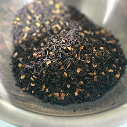 A close-up of loose-leaf tea, featuring dark black tea leaves mixed with small pieces of orange-brown citrus peel presented in a stainless steel bowl.