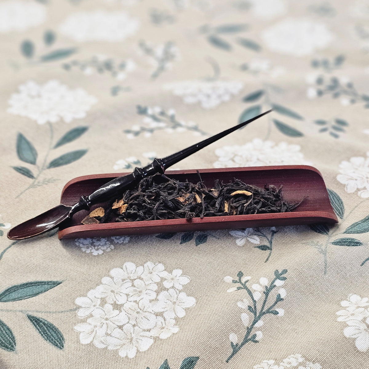 A dark wooden tea scoop resting on a reddish-brown rectangular dish filled with loose-leaf tea, placed on a beige fabric surface with a white floral print.