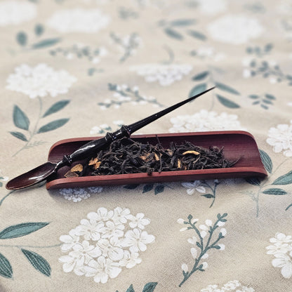A dark wooden tea scoop resting on a reddish-brown rectangular dish filled with loose-leaf tea, placed on a beige fabric surface with a white floral print.