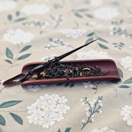 A dark wooden tea scoop resting on a reddish-brown rectangular dish filled with loose-leaf tea, placed on a beige fabric surface with a white floral print.