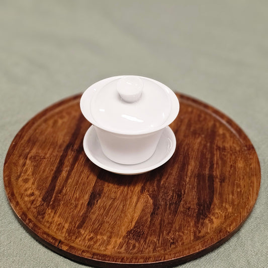 A white ceramic gaiwan (a bowl with a lid used for brewing tea) on a wooden platter.