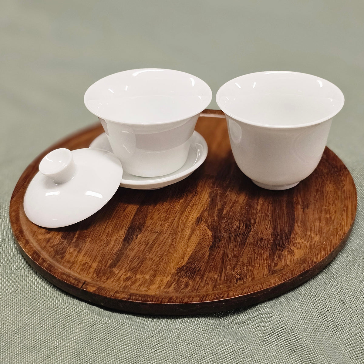 A white porcelain tea set with a miniature gaiwan with the lid resting on its saucer. Next to it is a teacup of similar shape and size. Both are displayed on a round wooden tray over a textured sage green fabric background.