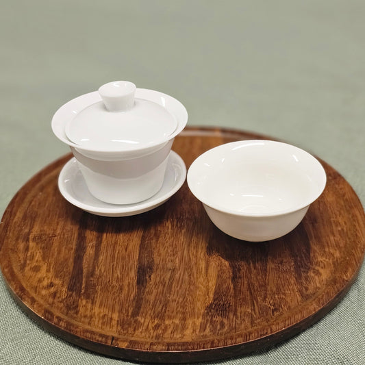 A white ceramic tea set consisting of a lidded gaiwan on a saucer and a matching teacup, arranged on a round wooden tray over a textured sage green fabric background.