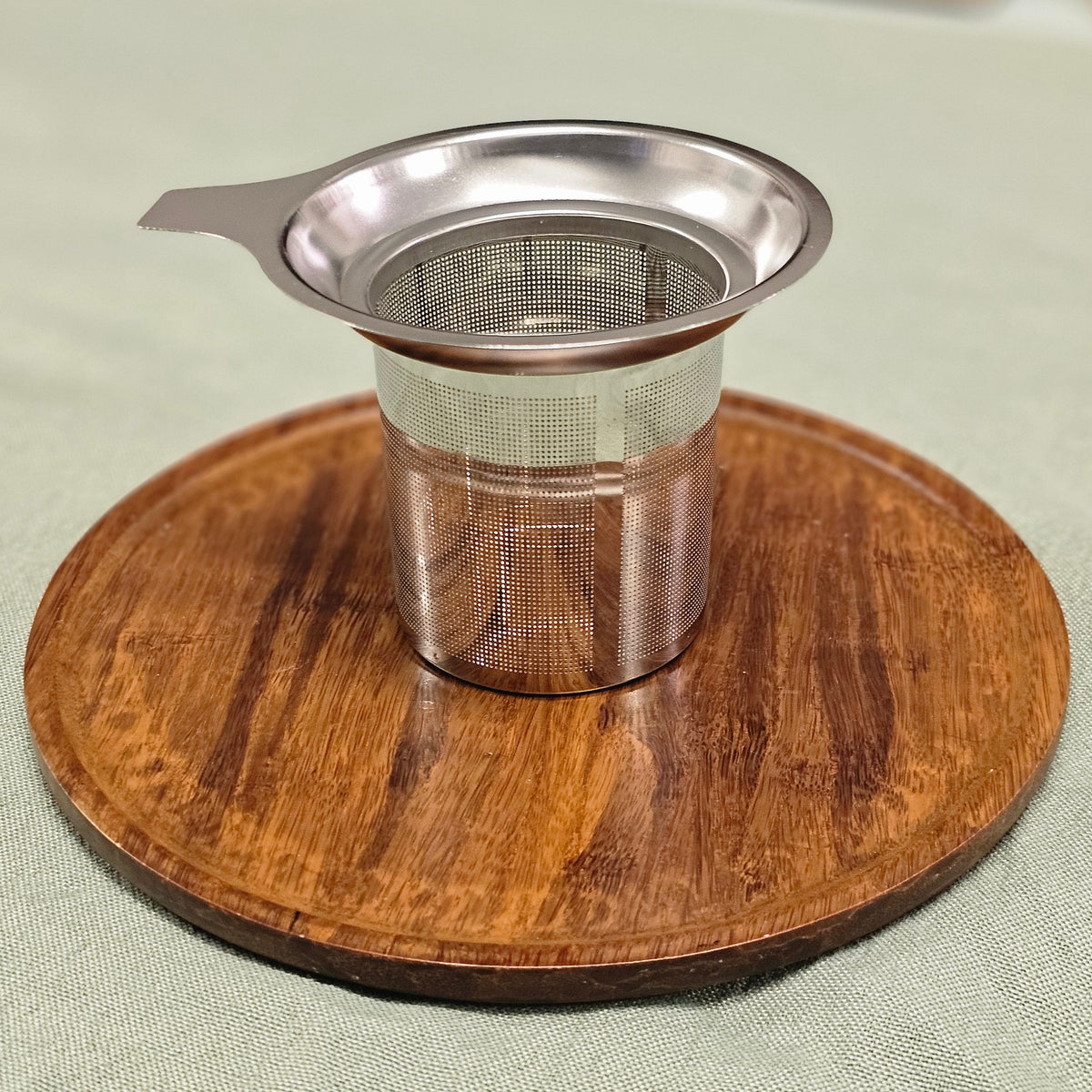 A stainless steel mesh tea infuser with a small handle, resting on a round wooden coaster over a light green fabric surface.