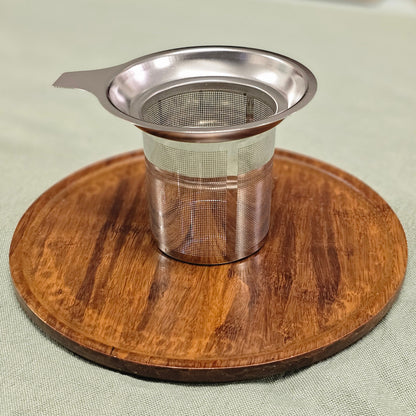 A stainless steel mesh tea infuser with a small handle, resting on a round wooden coaster over a light green fabric surface.