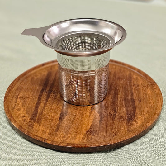 A stainless steel mesh tea infuser with a small handle, resting on a round wooden coaster over a light green fabric surface.